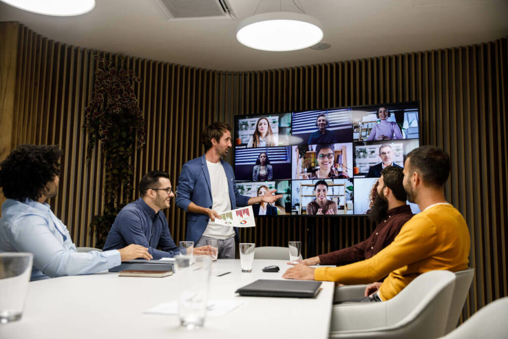 Employees in an office meeting room talking to employees working from home through a video call.